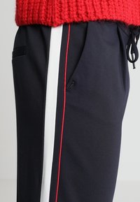 Jogging bleu marine avec rayures blanches sur les côtés et accents rouges, doté d'une ceinture élastique, d'un cordon de serrage et d'une poche latérale. Le matériau semble doux.