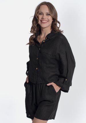 Donna con capelli ondulati sorridente, indossa una camicia nera ampia con bottoni e tasca e pantaloncini abbinati e comodi, mani nelle tasche, sfondo bianco.