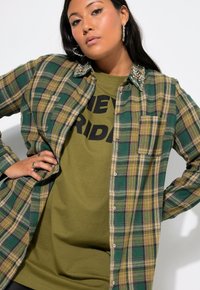 Groene en geruite button-up shirt met kralen op de kraag, gelaagd over een olijfgroen t-shirt met vetgedrukte zwarte tekst.