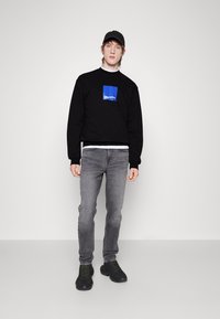 Felpa nera con una grafica a quadrato blu, jeans grigi e sneakers nere con dettagli verde. Il modello indossa anche un cappellino nero.