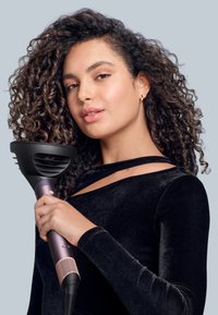 Hårfön med svart munstycke, lila handtag och rosa detaljer. Användaren håller den nära lockigt hår medan hon bär en svart sammetsöverdel.