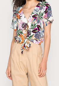 Woman wearing short-sleeve, colorful tropical print blouse tied at the waist with beige high-waisted pleated pants.