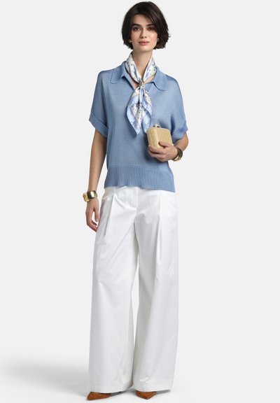 Femeie purtând un pulover cu mâneci scurte, bleu deschis, pantaloni largi albi, eșarfă florală, ținând o geantă plic mică bej și accesorii aurii.