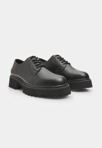 Chaussures noires en cuir avec un bout arrondi, un laçage et une semelle épaisse en caoutchouc noir texturé avec un relief prononcé.