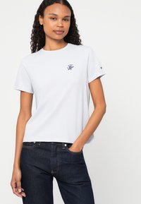 Balta kokvilnas T-krekls ar īsām piedurknēm, kuram uz krūtīm ir neliels izšūts logos tumši zilā krāsā un logotipa akcents uz piedurknes.