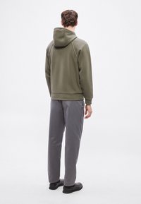 Oliwkowa zielona bluza z kapturem z tyłu oraz szare spodnie dresowe. Gładka tkanina o luźnym kroju i subtelnych detalach przeszyć. Czarne buty.