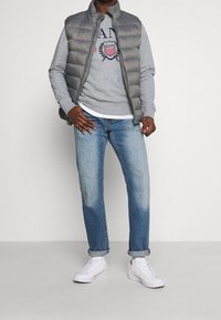 Chaleco de plumas gris sobre un sudadera gris clara, jeans azules con puños y zapatillas blancas. Los materiales son suaves y de corte relajado.