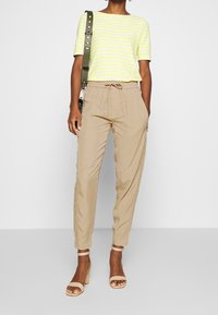 Gestreepte gele en witte top met korte mouwen, gecombineerd met beige tapering broeken. Het model draagt tan high-heeled sandalen en houdt een groene schouderband vast.