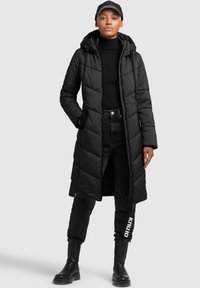 Mujer con un abrigo de plumas largo y negro con capucha, un cuello de tortuga negro, jeans negros con texto en la pierna, botas negras y una gorra de béisbol negra.