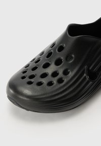 Black slip-on shoe made of textured plastic, featuring multiple circular cutouts and a curved shape, with a small logo accent.