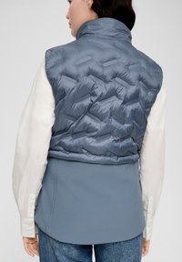 Gepolstertes blaues Weste mit hohem Kragen und einer strukturierten, glänzenden Oberfläche. Getragen über einem weißen Hemd, kombiniert mit blauen Stoffunterteilen und Jeans.
