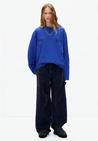 Bluza oversize albastră cu imprimeu "Stranger Things", combinată cu blugi largi de culoare închisă și bocanci verzi cu șireturi. Detaliu simplu cu lanțargintiu.