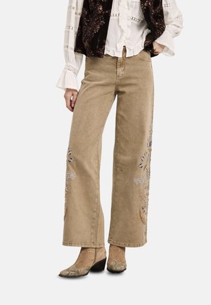 Mujer que lleva pantalones beige de pierna ancha con bordados florales, una blusa blanca con detalles de encaje, una chaqueta de terciopelo oscuro y botines de piel de serpiente.