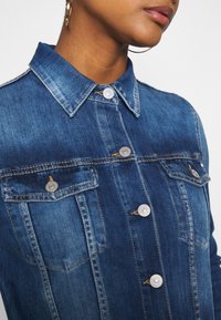 Mörkblå denimjacka med krage, silverknappar och två bröstfickor. Har detaljerad sömnad och en texturerad tyg.