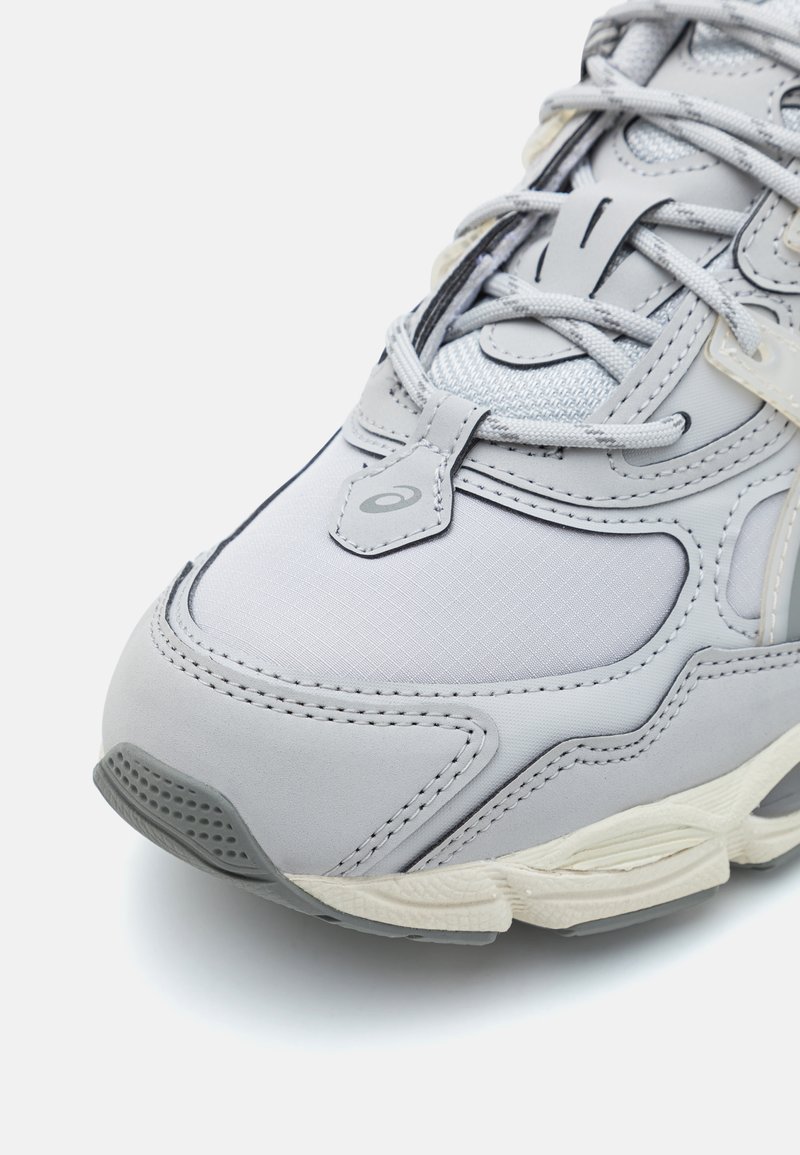 Grauer Sportschuh aus Mesh und synthetischem Material, mit abgerundetem Zehenbereich, strukturierten Akzenten und kontrastierender weißer Sohle. Verfügt über kunstvolle Nähte.