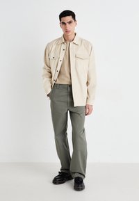 Chemise beige à boutons avec deux poches poitrine, t-shirt beige en dessous, pantalon vert olive et chaussures noires avec des coutures jaunes.