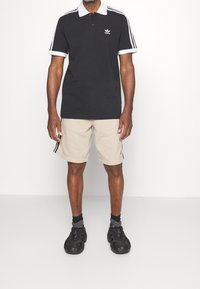 Man i svart Adidas-pikétröja med vita ränder och beige shorts med svarta sneakers som står mot en vit bakgrund.