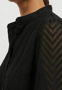Blusa nera con colletto alto, caratterizzata da un tessuto testurizzato e un motivo a zigzag sulle maniche. Include chiusure a bottoni al collo.