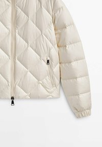 Veste matelassée beige avec coutures horizontales et en losange, fermeture éclair à l'avant, poche zippée et poignets élastiques sur la manche droite.