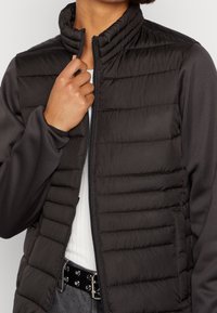 Gilet imbottito nero con colletto alto, texture trapuntata e tasche laterali. Tessuto liscio sulle maniche e chiusura con zip frontale.
