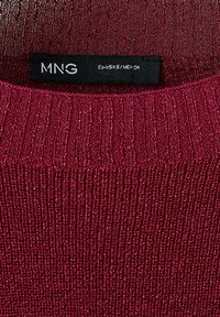 Étiquette de vêtement noire avec logo blanc MNG et taille EU-USA-S/MEX-CH sur un col de vêtement en maille côtelée rouge foncé.