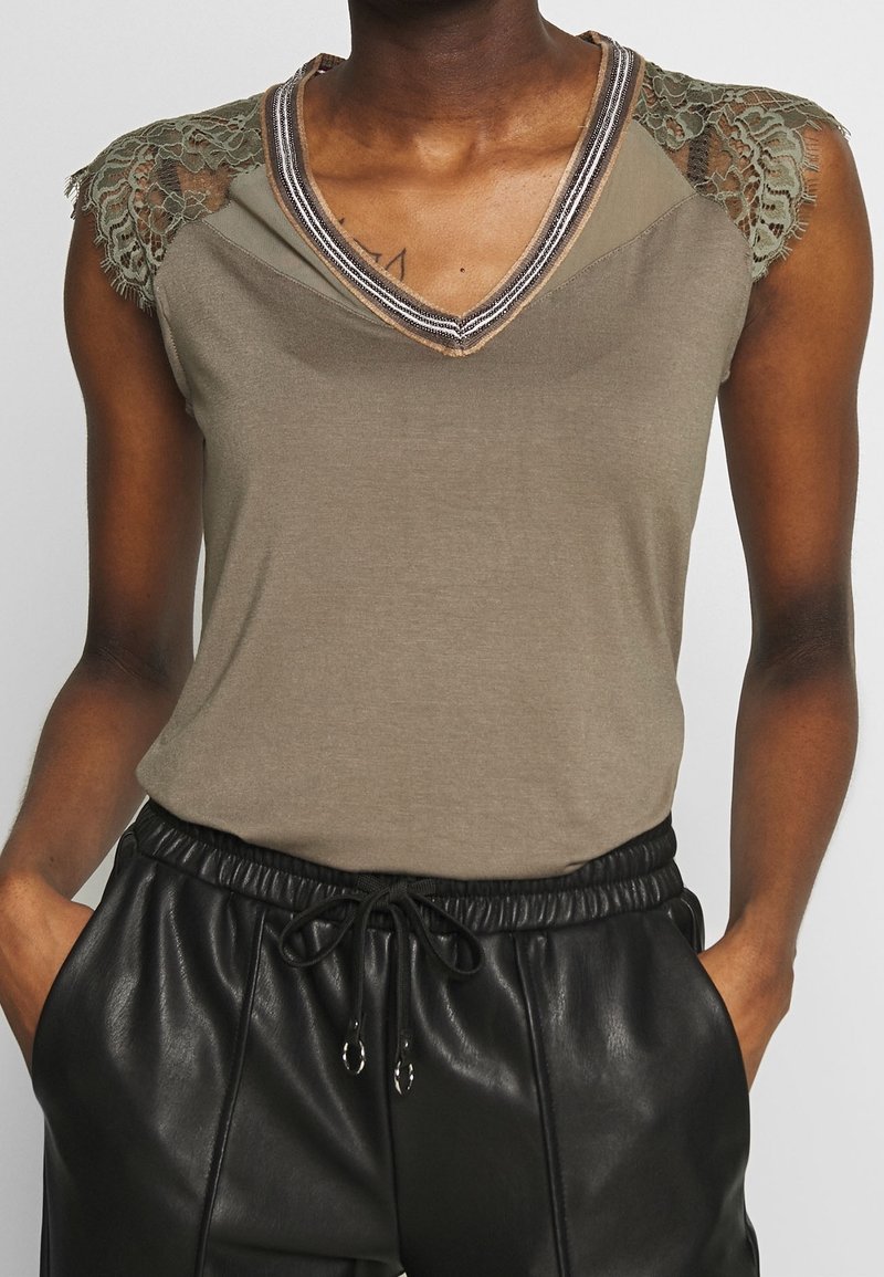 Olive green sleeveless top with lace shoulder accents, V-neckline, and decorative trim; paired with black faux leather pants.