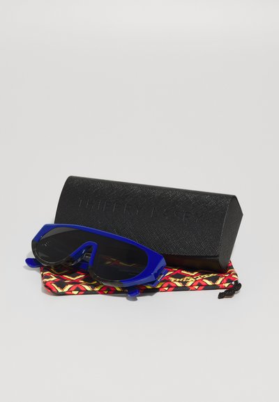Mėlynos saulės akinių rėmeliai su juodais objektyvais, kartu su juodu tekstūriniu dėklu ir spalvinga raštuota pakuote, kurioje dominuoja raudoni, oranžiniai ir geltoni akcentai.