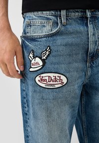 Muž v modrých džínách s dvěma nášivkami značky Von Dutch na levé stehně blízko kapsy a poutka na opasek.