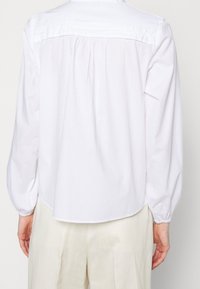 Valkoinen pitkähihaisen paita, jossa on kaareva helma, laskostusta takana ja joustavat hihansuut, valmistettu kevyestä kankaasta.