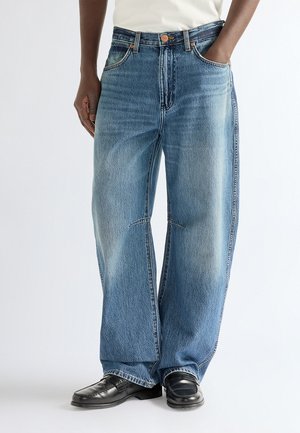Personne portant un jean bleu large et des chaussures en cuir noires, debout avec une main dans la poche, sur fond blanc.