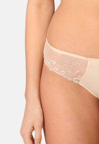 Beige underkläder med transparent meshdetaljering, som har intrikata blommönster broderade längs midjebandet och mjukt tyg på sidorna.