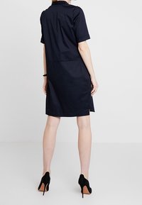 Donna che indossa un abito blu scuro a maniche corte e lunghezza ginocchio, con tacchi alti neri, in piedi di spalle contro uno sfondo chiaro e semplice.