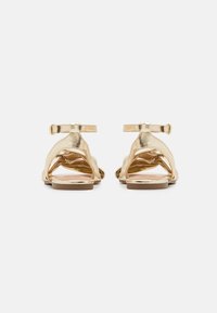 Sandales métalliques dorées avec une bride de cheville, un dos ouvert et un design croisillon sur le dessus. Semelle plate avec des accents matériels minimalistes.