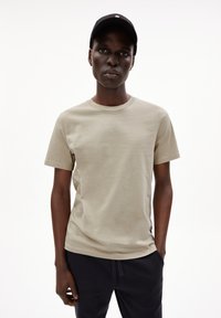 Licht beige katoenen t-shirt met een ronde hals, korte mouwen en een eenvoudige ontwerpen. In combinatie met donkerblauwe broek en een zwarte pet.