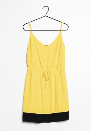 Robe sans manches jaune avec un décolleté en V, taille élastique, liens ajustables et ourlet noir. Texture lisse, suspendue à un crochet noir.