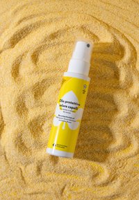 Olio per capelli protettivo dal sole in una bottiglia cilindrica bianca con etichetta gialla, caratterizzata da grafiche di foglie verdi e un erogatore spray, posata sulla sabbia gialla.