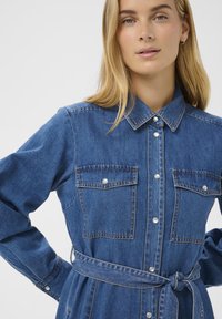 Denim shirt jurk in medium blauw met een knoopsluiting aan de voorkant, twee borstzakken en een strikceintuur in de taille. Heeft een klassieke kraag en lange mouwen.