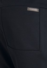 Zwart textiel met een gladde textuur. Een rechthoekige zak aan de zijkant en een klein label met "Haldessari" dicht bij de naad van de zak.