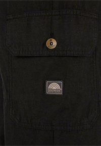 Sort stofskjorte med en stor lomme foran med en messingknap. Indeholder et rektangulært logo-patch med teksten "SOUTHPOL" på lommen.