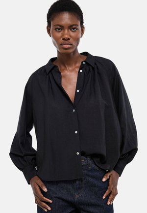 Femme aux cheveux courts portant une chemise noire ample à boutons et un jean bleu foncé, debout les mains dans les poches devant un fond uni.