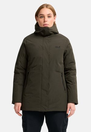 Persoon die een donkergroene winterjas met capuchon draagt en ontspannen met de handen langs het lichaam staat tegen een effen achtergrond.