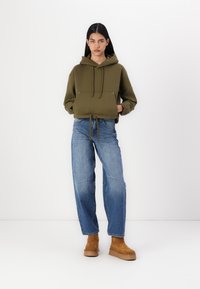 Ελαιοπράσινη cropped hoodie με κορδόνι, συνδυασμένη με ψηλόμεσα μπλε τζιν και καφέ σουέτ μποτάκια μέχρι τον αστράγαλο σε λευκό φόντο.
