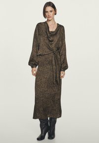 Donna in piedi che indossa un abito midi a maniche lunghe, marrone a fantasia, con vita legata, abbinato a stivali neri in pelle al ginocchio.