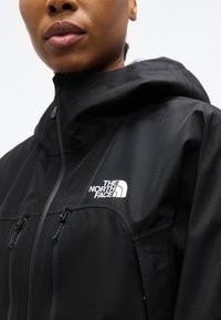 Gros plan sur une personne portant une veste noire The North Face avec des poches poitrine zippées et un col relevé.