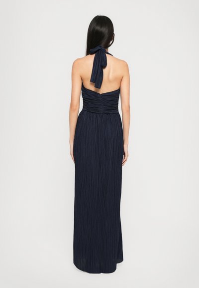 Long, navy dress with a halter neck and tied back. Features ruched bodice and flowing, textured fabric. Elegant floor-length design.