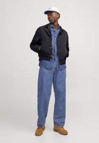 Giubbotto bomber nero su una camicia di jeans azzurro chiaro, abbinato a jeans blu larghi e stivali marroni, completato da un berretto crema.