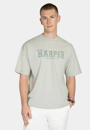 Jonge man met kort haar, gekleed in een lichtgrijs oversized T-shirt met de tekst "HARPER", witte broek, een horloge en polsbandjes, staand tegen een lichte achtergrond.