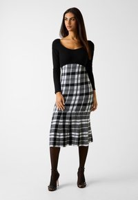 Schwarzes langärmliges Kleid mit einem gerippten Oberteil und einem karorierten Rock in Grautönen und Weiß, ausgestattet mit einem ausgestellten Saum und Falten.