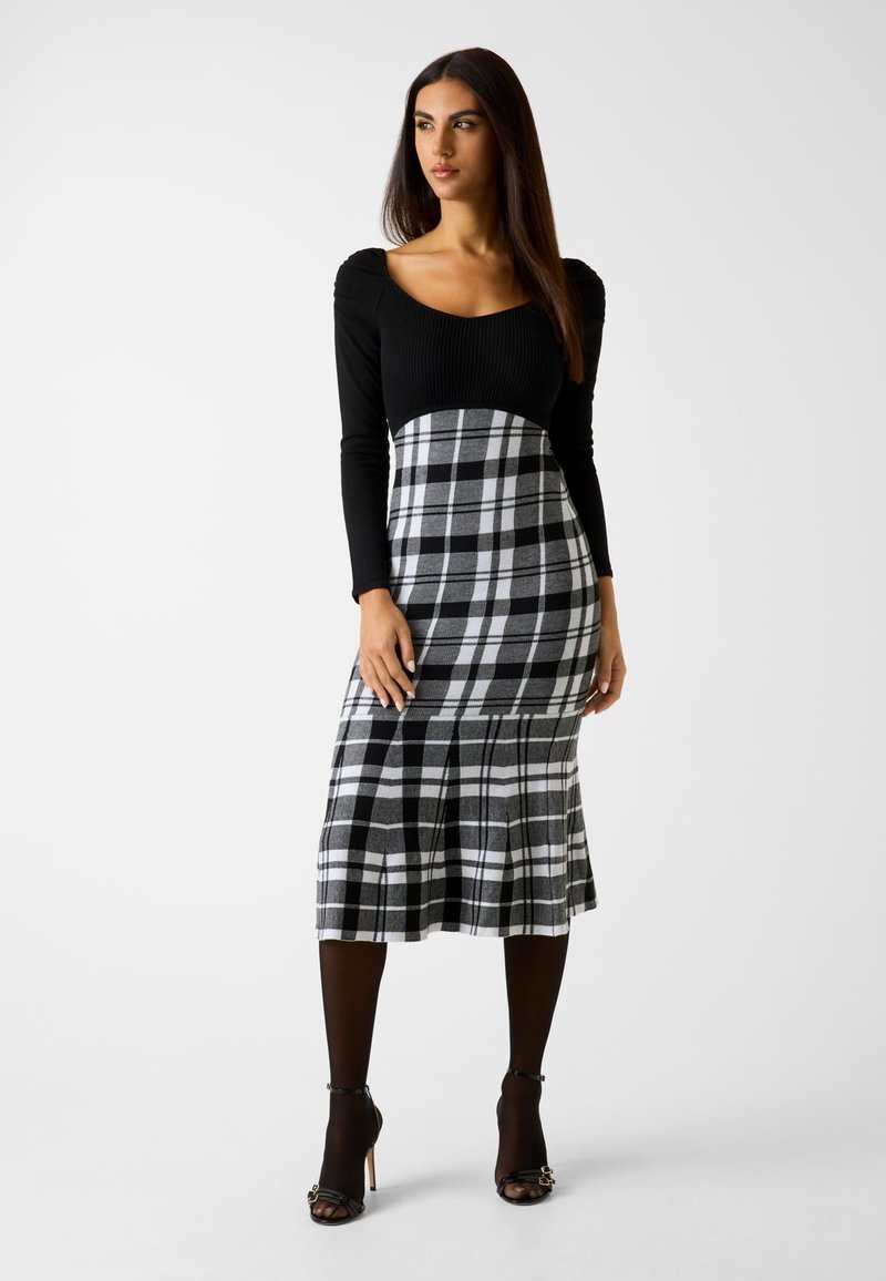 Schwarzes langärmliges Kleid mit einem gerippten Oberteil und einem karorierten Rock in Grautönen und Weiß, ausgestattet mit einem ausgestellten Saum und Falten.