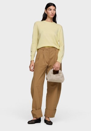 Donna che indossa un maglione giallo chiaro, pantaloni marroni con ampio risvolto, ballerine nere in rete, che tiene una piccola borsa beige pelosa, in piedi su uno sfondo bianco.